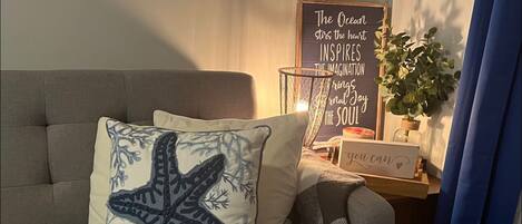 A cozy living room with a grey sofa, a starfish pillow, a checkered throw blanket, a wall-mounted sign with inspirational quotes, and a potted plant.