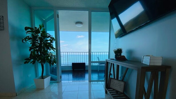 A room with a balcony, a potted plant, a TV mounted on the wall, and a small table with a plant on it. - Condo with Amazing Ocean View in Fajardo near Villa Marina (Fajardo)