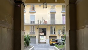 Un couloir en arc de cercle mène à un bâtiment à la façade jaune, une voiture jaune est garée devant, et l’entrée est bien visible.