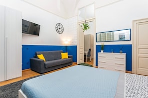 Un dormitorio con cama, un sofá gris con cojines amarillos, cómoda, espejo, reloj y televisor en la pared.