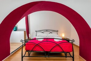 Una cama con colcha roja y almohadas blancas, una lámpara de noche y un arco rojo que enmarca la habitación.