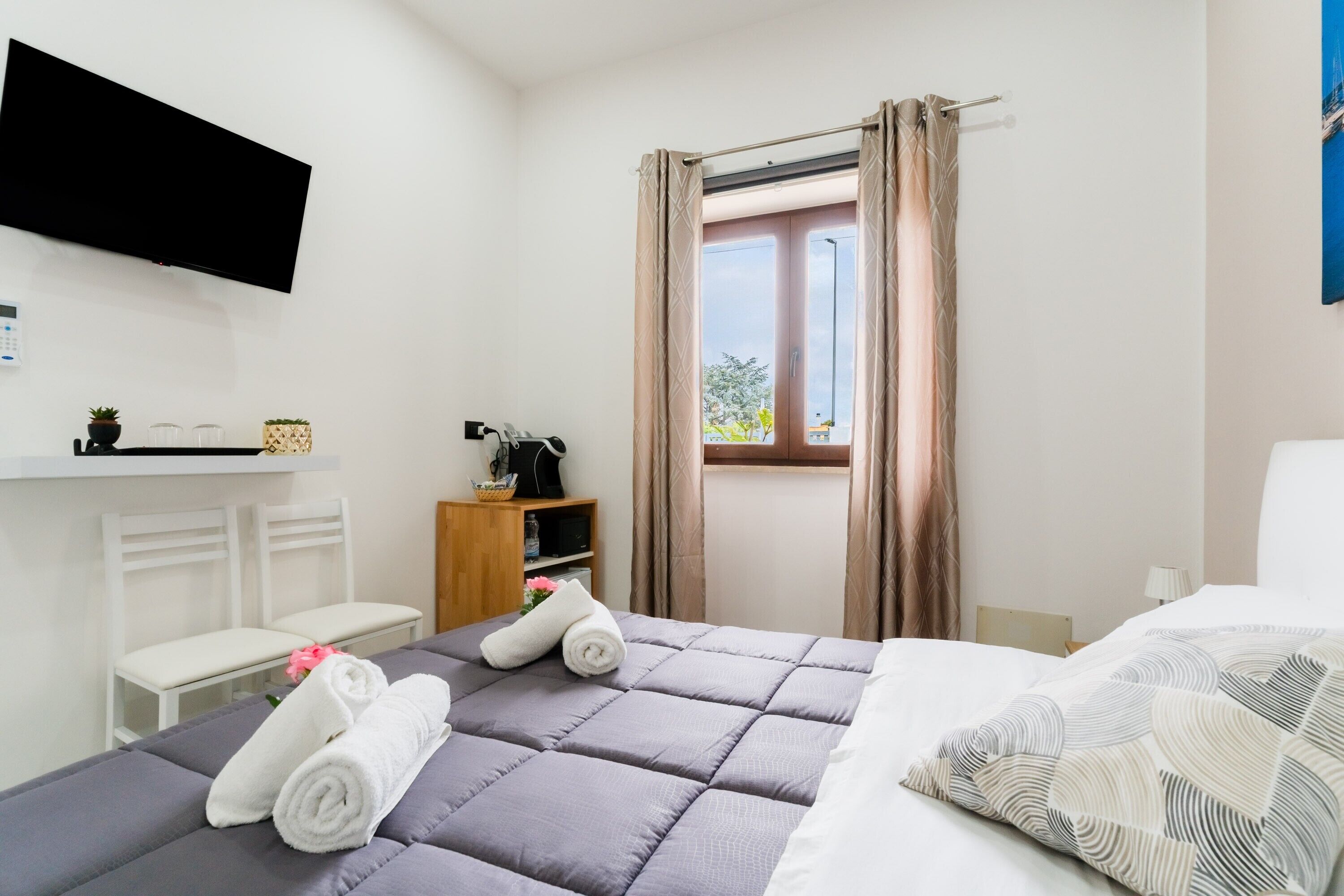 Una habitación de hotel con una cama, televisor montado en la pared, ventana con cortinas, un escritorio pequeño con lámpara y estante con adornos.