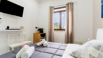 Una habitación de hotel con una cama, televisor montado en la pared, ventana con cortinas, un escritorio pequeño con lámpara y estante con adornos.