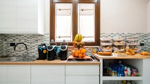 Una cocina con un lavamanos, dos cafeteras, una frutera y diversos alimentos sobre la encimera.