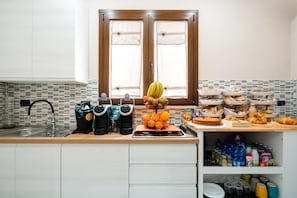 Una cocina con un lavamanos, dos cafeteras, una frutera y diversos alimentos sobre la encimera.
