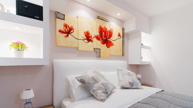 Une chambre à coucher moderne avec un lit, des oreillers, une table de chevet, une étagère murale avec un vase et un tableau.