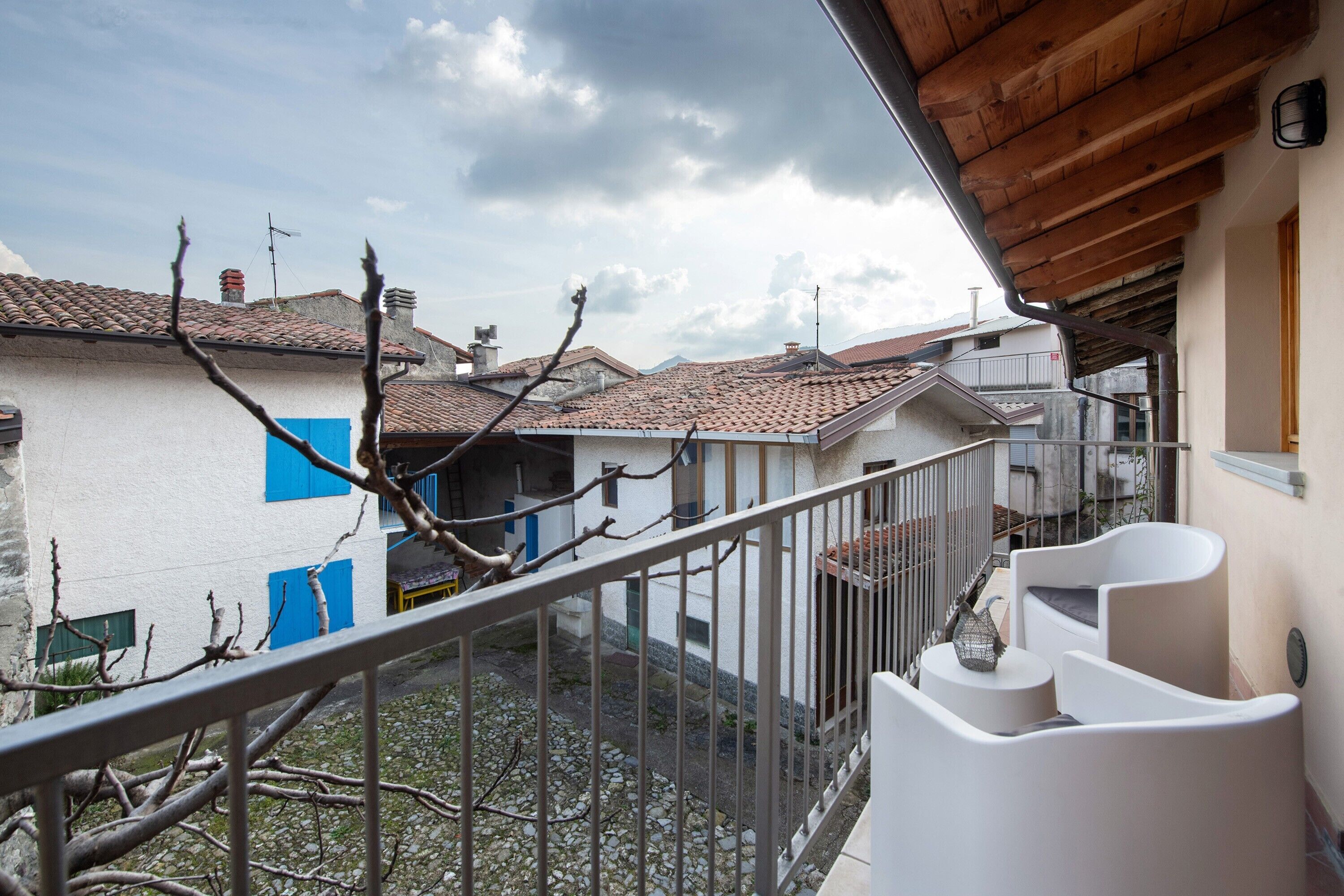 Un balcón con barandilla metálica, una pared blanca con ventana azul y un techo de tejas al fondo.