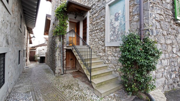 Une ruelle étroite pavée, bordée d’un bâtiment en pierre doté d’une porte en bois, d’un escalier avec des rampes en métal et d’un arbuste aux feuilles vertes.
