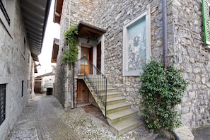A narrow cobblestone street with a stone building featuring a wooden door, a staircase with metal railings, and a green-leaved bush.