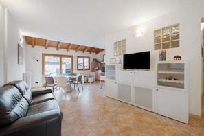 A modern living room with a leather sofa, a dining area with chairs, a kitchen with cabinets, and a TV mounted on the wall.