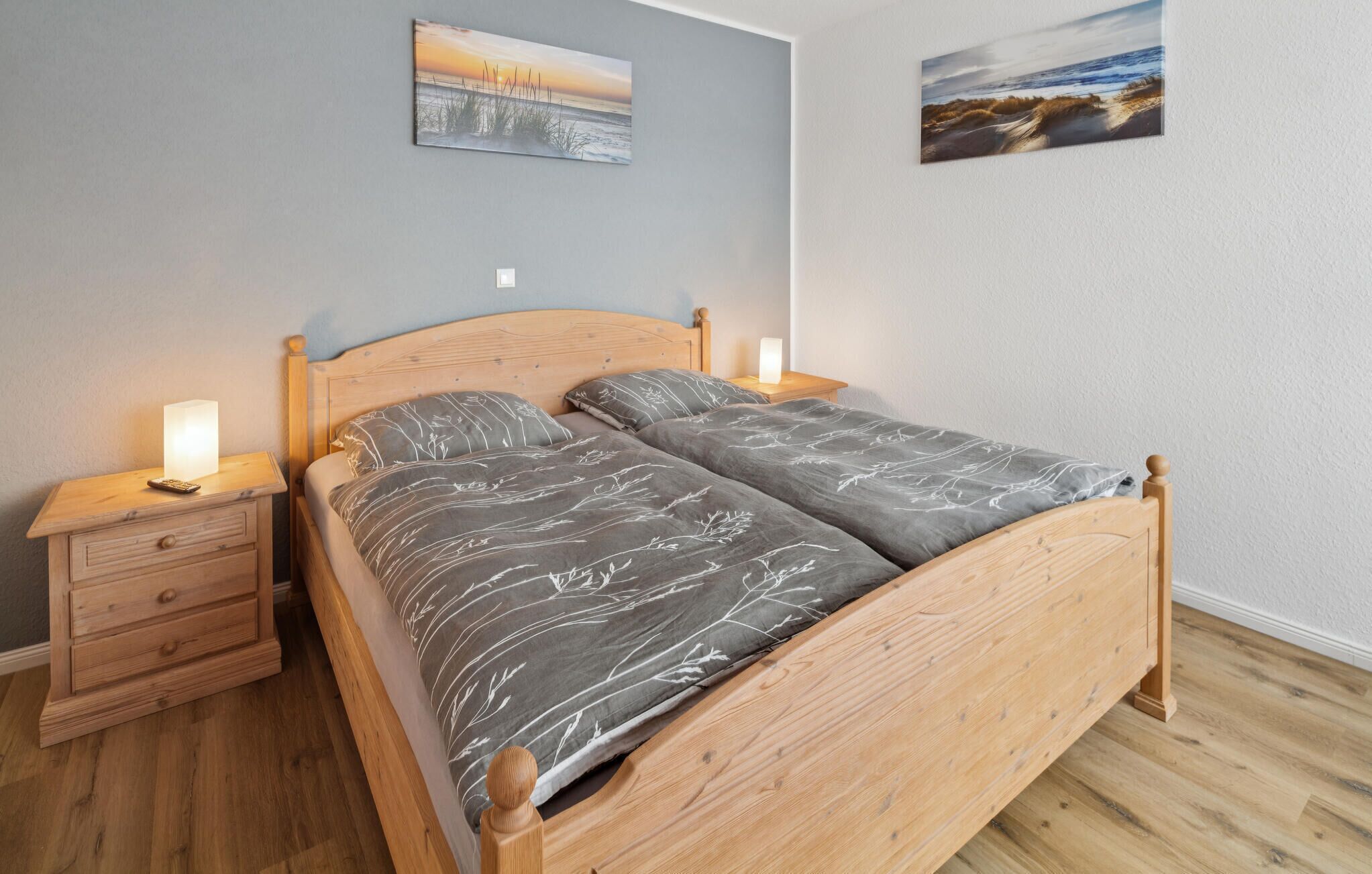Een houten bed met een grijs geborduurde spreideken, twee nachtkastjes met lampen, en twee aan de muur hangende schilderijen.