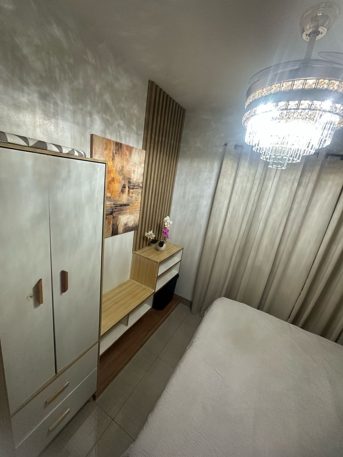 A hotel room with a bed, a wardrobe, a vanity with a mirror, a chandelier, and a painting on the wall.