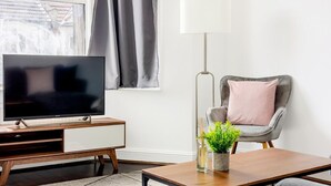 Ein modernes Wohnzimmer mit Flachbildfernseher auf einem Holz-TV-Möbel, einem grauen Sessel mit einem rosafarbenen Kissen, einer Stehlampe und einem kleinen Tisch mit einer Pflanze.