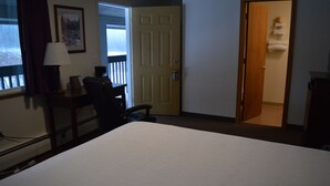 Ett hotellrum med en säng, ett skrivbord med en lampa, en stol, en dörr och utsikt över omgivningen.