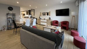A modern living room with a sofa, red chairs, a dining area, and a kitchen in the background. - Charming single-story villa with 3 bedrooms, tastefully decorated, beautiful exterior. (CANET PLAGE)