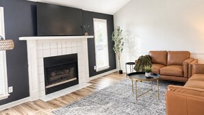 A living room with a fireplace, a ceiling fan, a brown leather sofa, and a patterned rug. - Juniper: 3 bedroom w/ pool (Martinez)