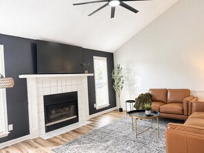 A living room with a fireplace, a ceiling fan, a brown leather sofa, and a patterned rug. - Juniper: 3 bedroom w/ pool (Martinez)