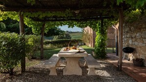 A stone table with picnic supplies under a pergola with vines, surrounded by gravel and greenery. - Villa Charming on the Hills | Mixed of Authentic and Modern Tuscan Style (Siena)