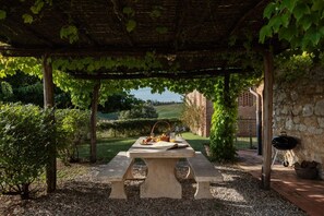 A stone table with picnic supplies under a pergola with vines, surrounded by gravel and greenery. - Villa Charming on the Hills | Mixed of Authentic and Modern Tuscan Style (Siena)