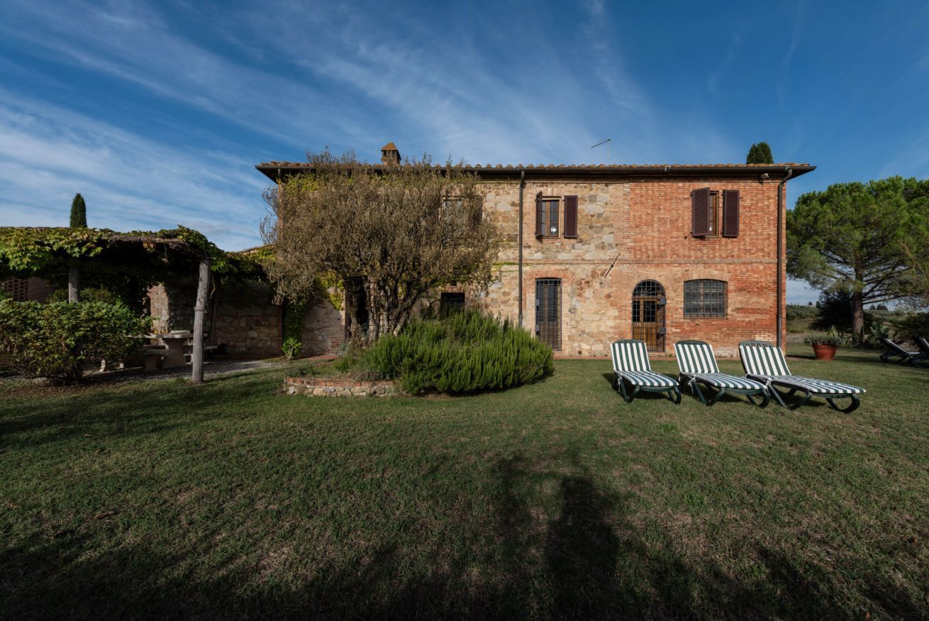 A rustic stone building with a tiled roof, a small garden with a lawn and four striped lounge chairs.