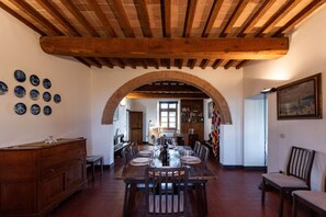 A dining area with a wooden table set for six, surrounded by chairs. An arched brick structure spans the room, and a wooden ceiling beam is visible.