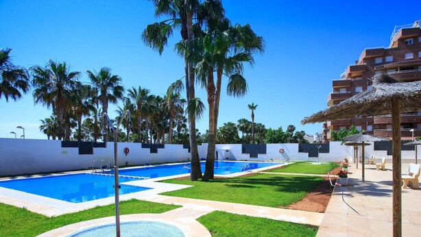 A swimming pool area with a circular mosaic design, palm trees, and a building with a thatched roof. - Las Terrazas I by Interhome (Oropesa del Mar)