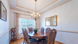Une salle à manger avec une table en bois, six chaises, un lustre, un miroir et une fenêtre avec des stores.