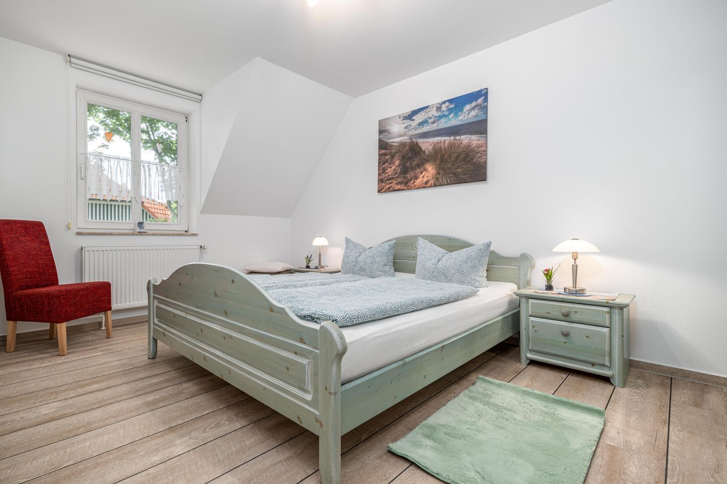 Un dormitorio con cama, dos mesitas de noche con lámparas, una silla, una ventana con vistas y un cuadro en la pared.
