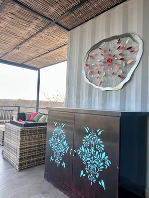 Een overdekt terras met een rieten bank en kussens, een decoratief metalen object aan de muur, en een donker houten kast met bloemmotieven.