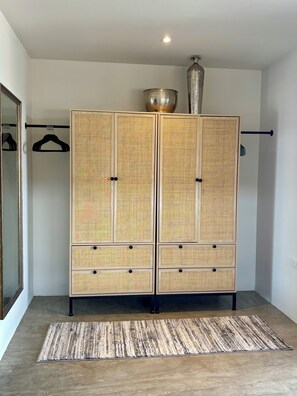 A room with a large, two-tiered rattan wardrobe, a hanging rack, a mirror, and a textured rug on a tiled floor.
