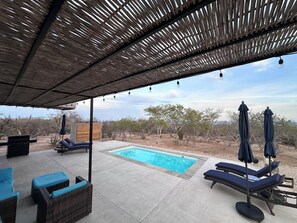 Uma área na beira da piscina com espreguiçadeiras e piscina, cercada por uma pérgula de madeira e paisagem desértica.