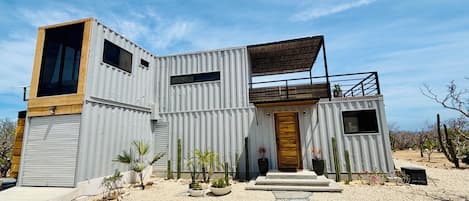 A modern, container-style house with a large garage, a wooden front door, and a balcony.
