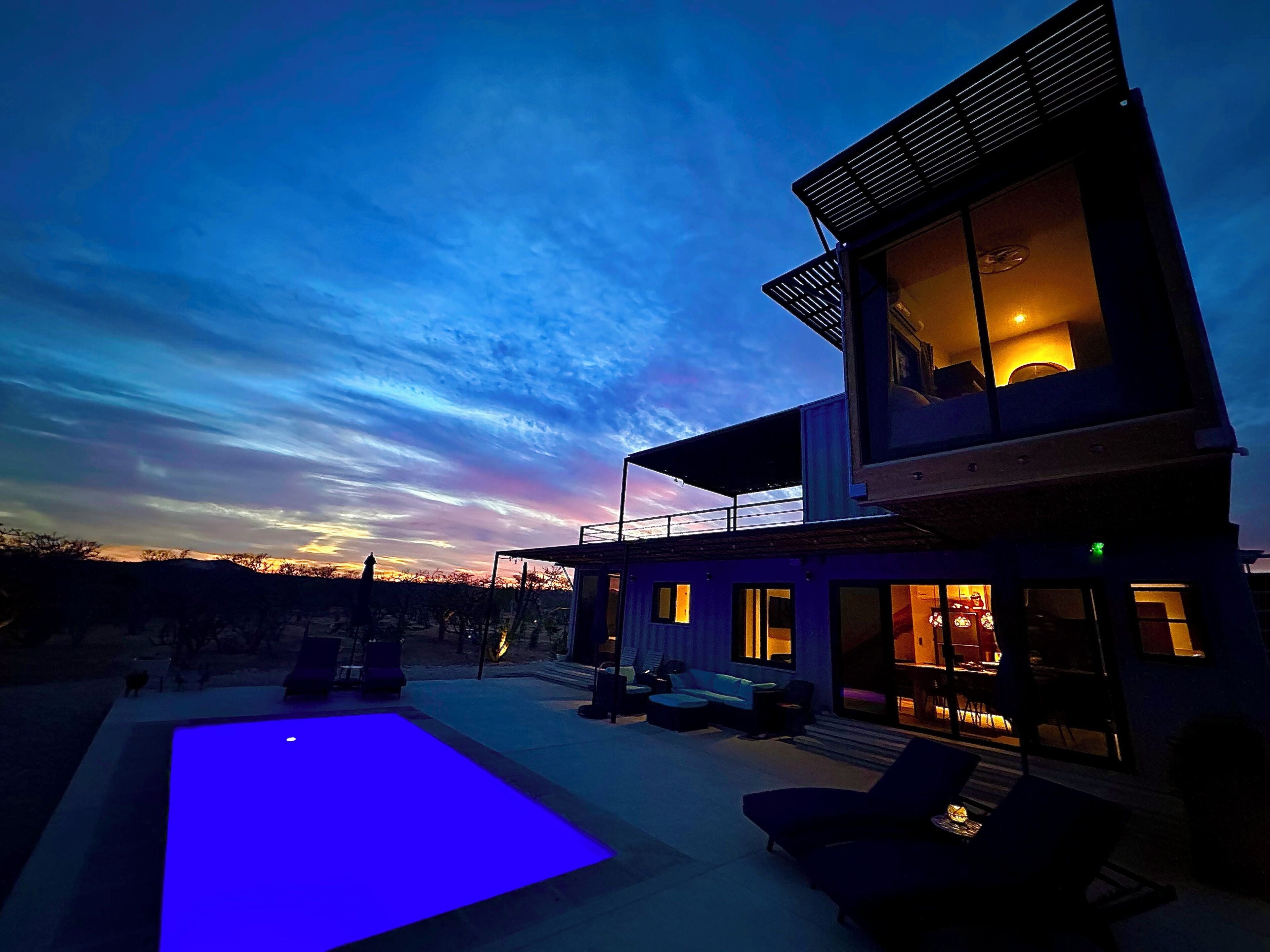 Una casa moderna de dos pisos con piscina, mobiliario exterior y vista al atardecer.