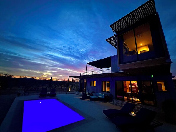 A modern two-story house with a pool, outdoor seating, and a view of the sunset.