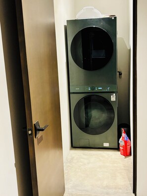 Uma porta que dá para uma lavanderia com máquina de lavar e secadora empilhadas, um painel de controle e um saco de detergente no chão.