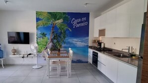 Eine moderne Küche mit weißen Schränken, einem Essbereich mit Tisch und Stühlen, einem großen Wasserspender und einem an der Wand befestigten Fernseher.