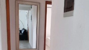Ein schmaler Flur mit weißen Wänden und gefliestem Boden. Eine Tür mit Spiegel ist an der Wand zu sehen, die einen Raum mit einem Bett und einem Fenster reflektiert.