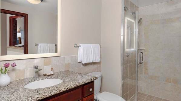 Un baño con lavamanos de encimera de granito, un espejo, toallero y ducha con mampara de vidrio.
