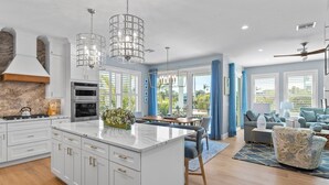A modern kitchen with white cabinetry, a central island, and pendant lighting. There is a dining area with a blue dining set and a view of the outdoors. - Salt Dreams: 1 Block to Beach! Pool, Spa, Games, Rooftop Deck by Anna Maria Life Vacation Rentals (Anna Maria)