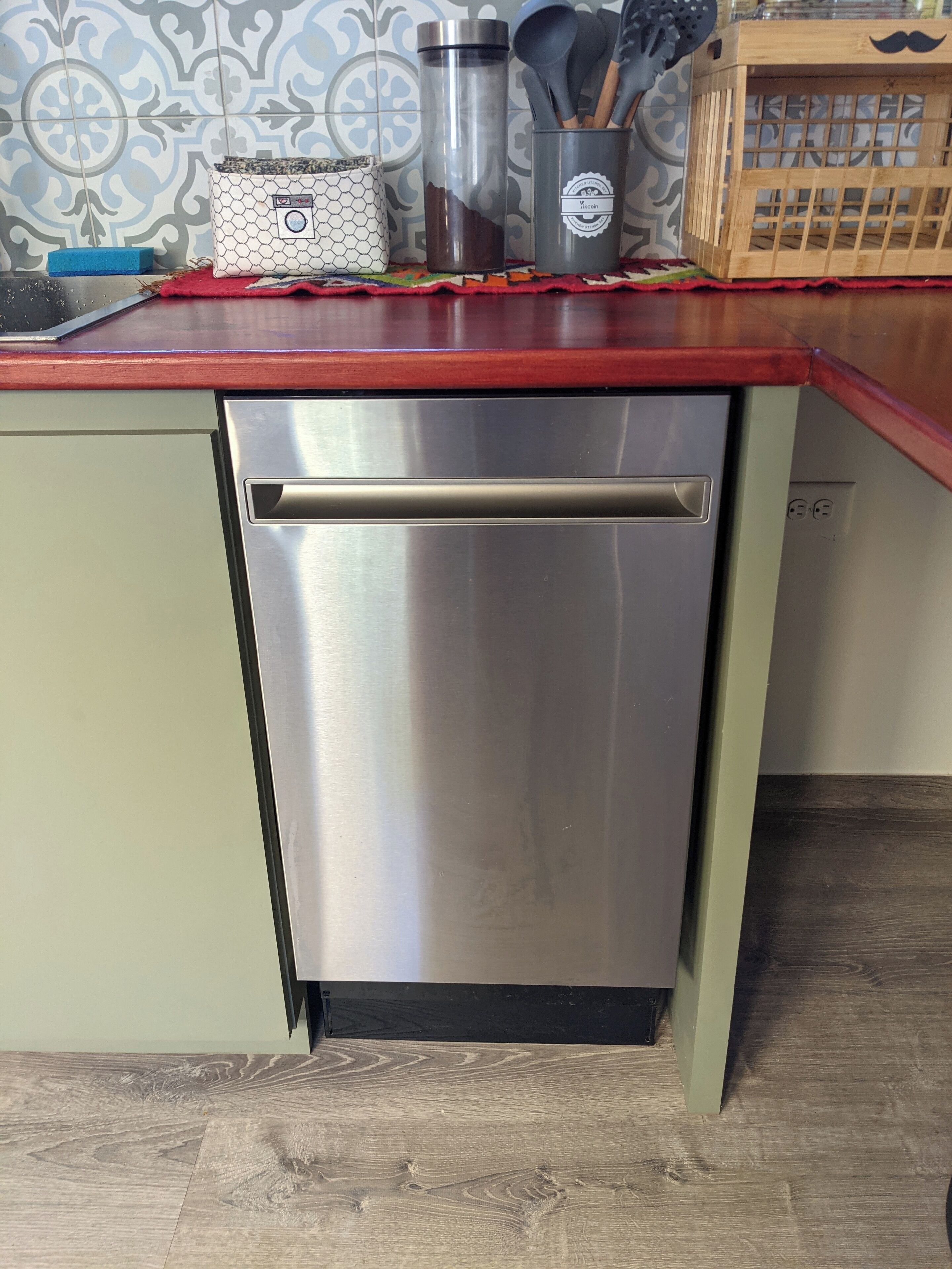 A stainless steel dishwasher in a kitchen with a red countertop, a hexagonal patterned soap dish, and a wooden crate with a black mustache design.