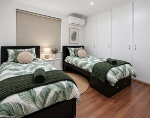 A bedroom with two beds, a bedside table with a lamp, a wall-mounted air conditioner, and a large wardrobe. - Home Homly | Cozy Family Getaway (Rockingham)