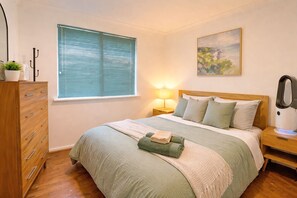 A bedroom with a bed, a nightstand, a lamp, a dresser, a window with blinds, and a painting on the wall. - Home Homly | Cozy Family Getaway (Rockingham)