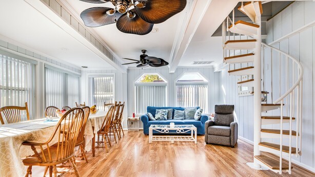 Amplio salón con sofá azul, mesa de comedor de madera y sillas. Se ve una escalera con barandilla blanca a la derecha.