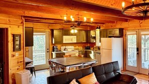 A cabin interior with a wooden ceiling, a dining area, a kitchen with a refrigerator, and a black leather sofa with pillows.