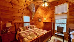 A cozy wooden cabin bedroom with a bed, a chair, a nightstand, a dresser, a ceiling fan, and two windows.