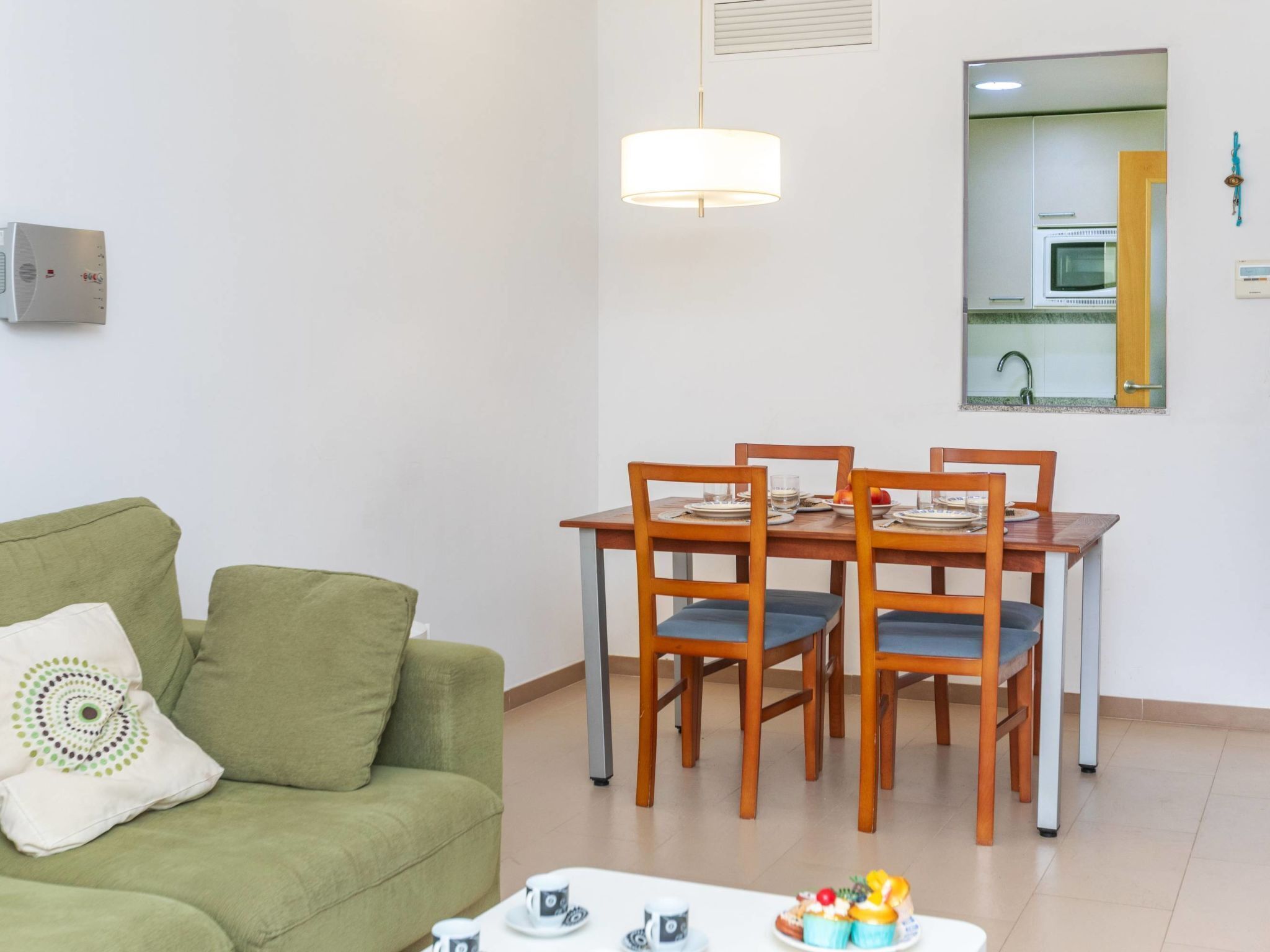 A living room with a green sofa, a dining table with chairs, and a kitchen visible through an open door.