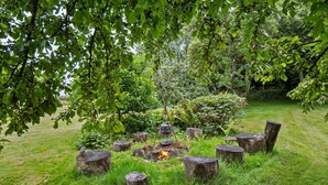 Un feu de camp entouré de souches d’arbres, dans une zone herbeuse parsemée d’arbres et d’arbustes.