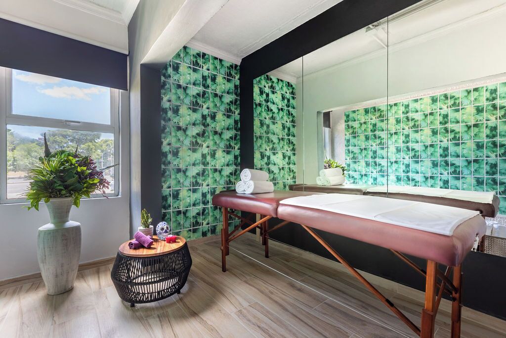 Ein Spa-Raum mit Holzboden, einer Massageliege, einem kleinen runden Tisch mit Pflanze und einem Fenster mit Blick auf Grünflächen.