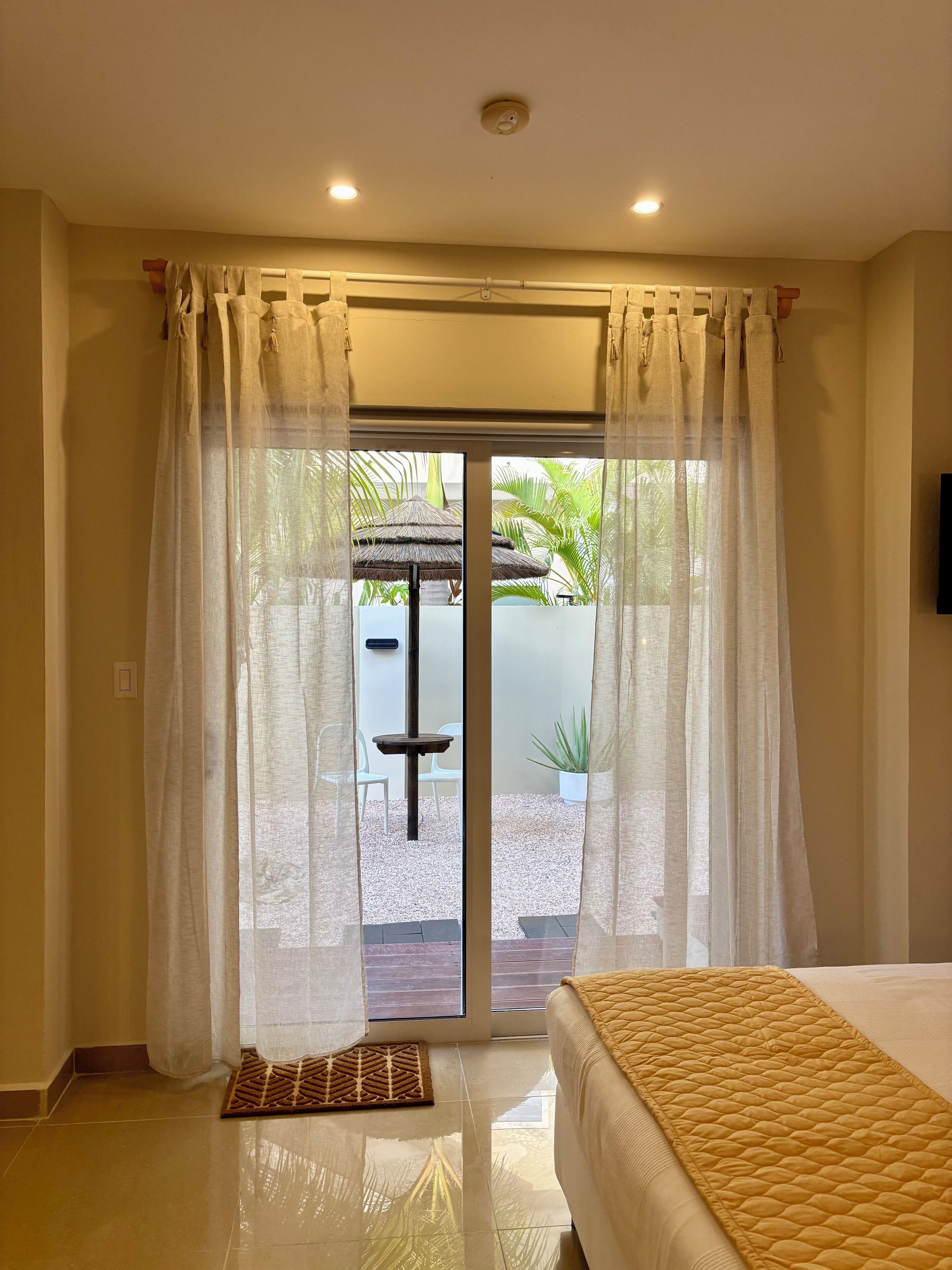 A hotel room with a bed, a small table, and a view of an outdoor area with a palm tree.