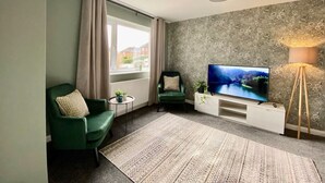 Ein modernes Wohnzimmer mit einem Flachbildfernseher, zwei grünen Sesseln, einem kleinen Beistelltisch, einer Stehlampe und einem gemusterten Teppich.